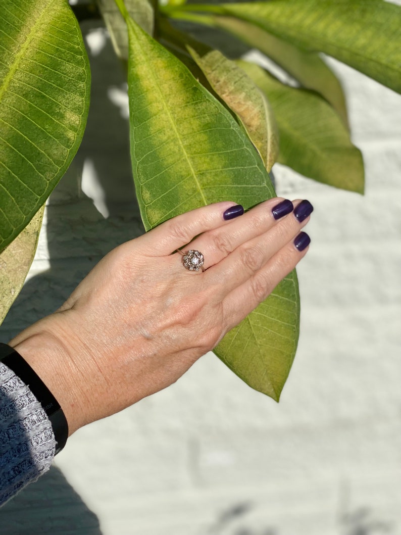 Puede incluir: Un anillo de oro con una piedra grande, redonda y transparente rodeada de piedras m&aacute;s peque&ntilde;as. El anillo est&aacute; en el dedo de una persona.