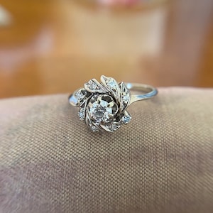 Puede incluir: Un anillo de plata con un gran diamante redondo rodeado de diamantes m&aacute;s peque&ntilde;os en un dise&ntilde;o floral.