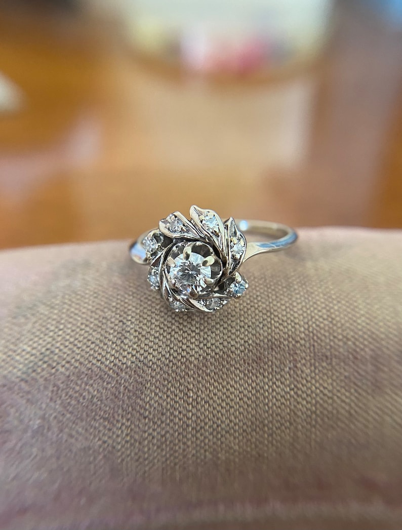 Puede incluir: Un anillo de plata con un gran diamante redondo rodeado de diamantes m&aacute;s peque&ntilde;os en un dise&ntilde;o floral.