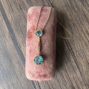 Op de afbeelding: Een gouden ketting met een hanger met twee ronde blauwe edelstenen. De ketting wordt gepresenteerd in een roze fluwelen doosje.