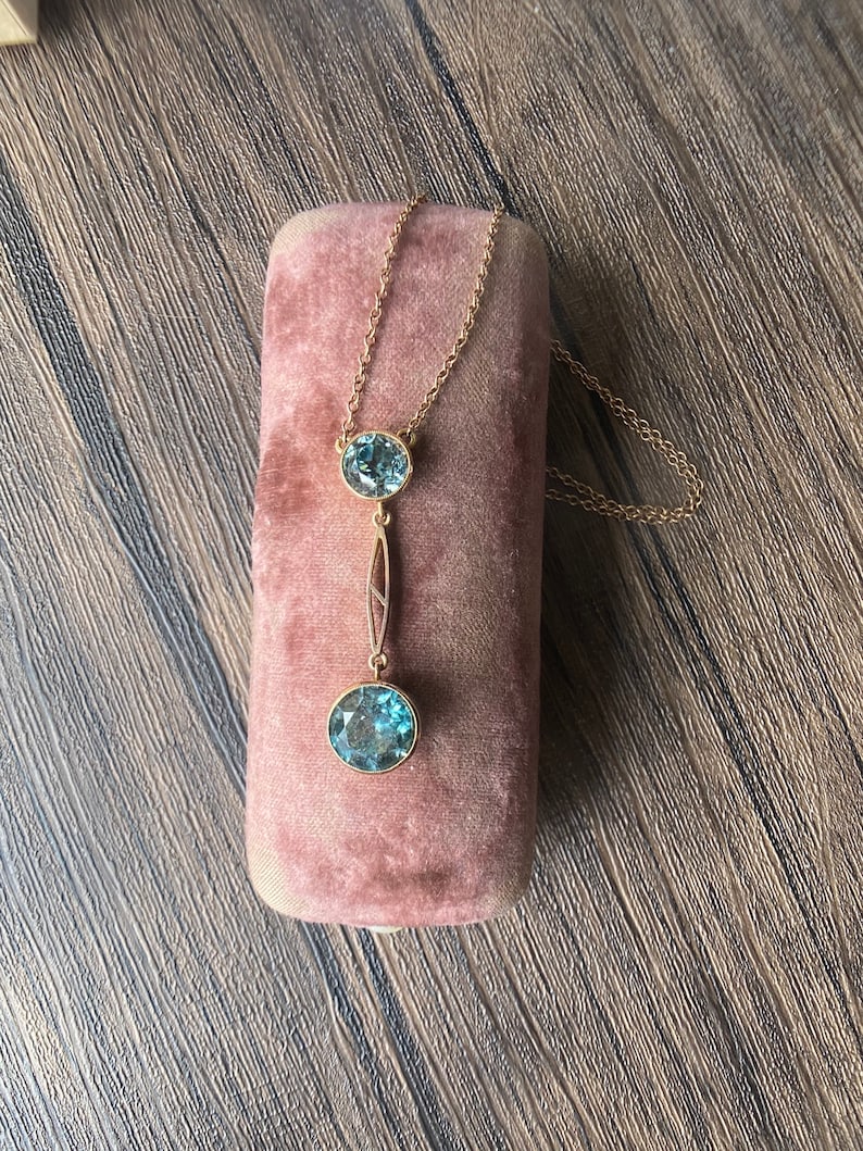 Op de afbeelding: Een gouden ketting met een hanger met twee ronde blauwe edelstenen. De ketting wordt gepresenteerd in een roze fluwelen doosje.