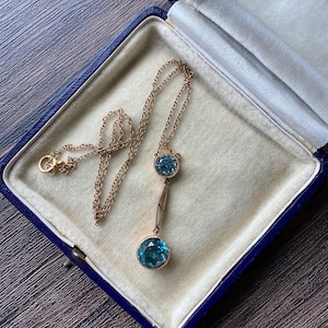 Op de afbeelding: Een gouden ketting met een druppelhanger met twee ronde blauwe edelstenen. De ketting ligt in een blauwe fluwelen juwelendoos.