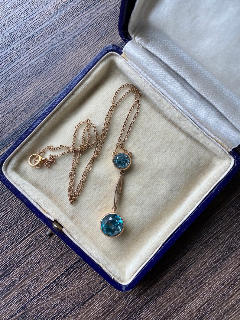 Op de afbeelding: Een gouden ketting met een druppelhanger met twee ronde blauwe edelstenen. De ketting ligt in een blauwe fluwelen juwelendoos.