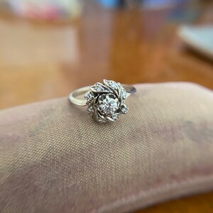 Puede incluir: Un anillo de plata con un diamante redondo central rodeado de diamantes m&aacute;s peque&ntilde;os en un dise&ntilde;o floral.
