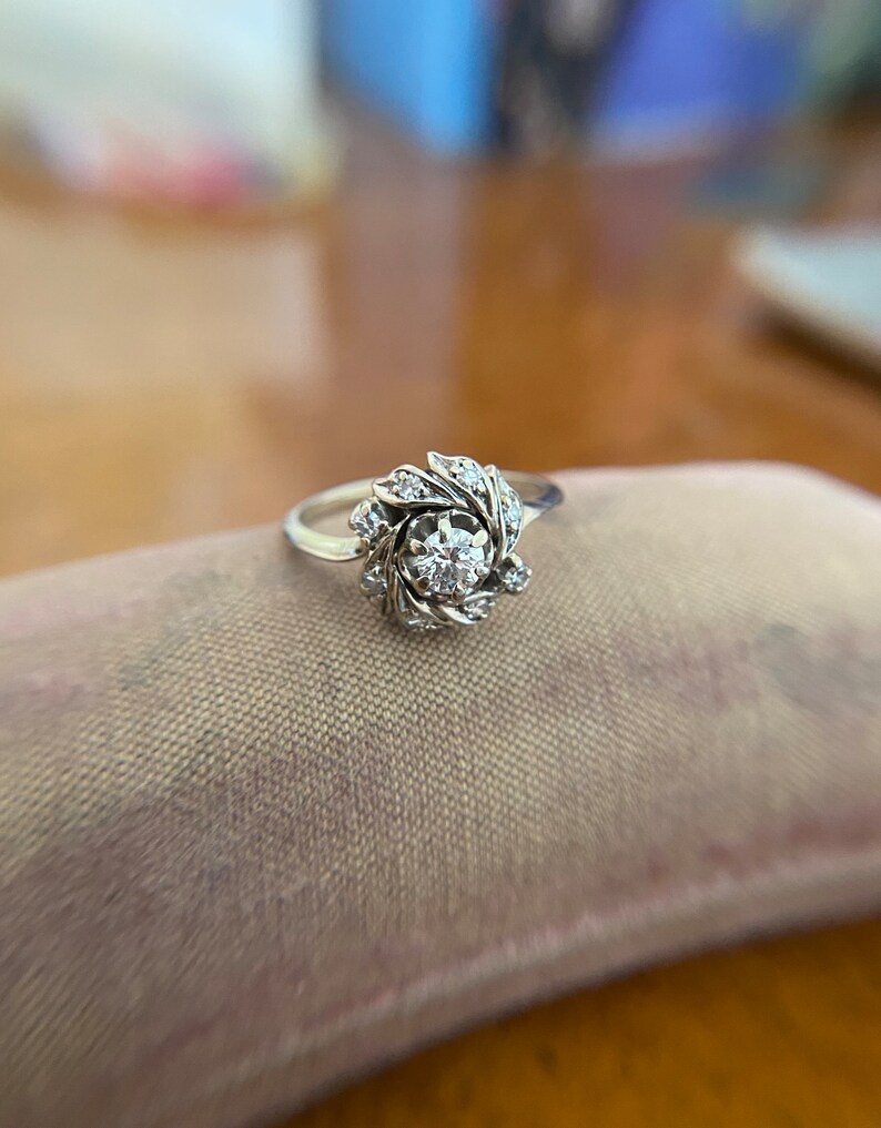 Puede incluir: Un anillo de plata con un diamante redondo central rodeado de diamantes m&aacute;s peque&ntilde;os en un dise&ntilde;o floral.