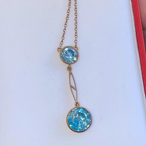 Op de afbeelding: Een gouden ketting met twee blauwe edelstenen. De ketting heeft een fijne ketting en een druppelvormig ontwerp met een grotere edelsteen aan de onderkant.