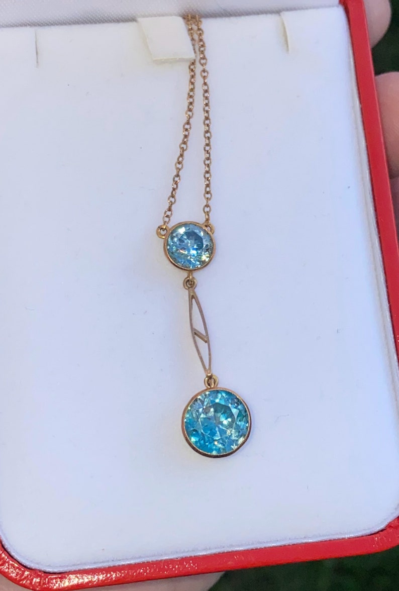 Op de afbeelding: Een gouden ketting met twee blauwe edelstenen. De ketting heeft een fijne ketting en een druppelvormig ontwerp met een grotere edelsteen aan de onderkant.