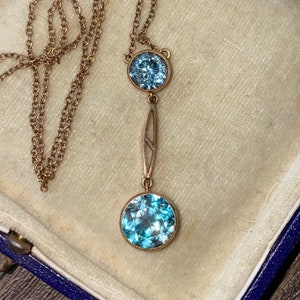 Op de afbeelding: Een gouden ketting met een hanger met twee ronde, lichtblauwe edelstenen. De hanger heeft een delicaat art deco-ontwerp.