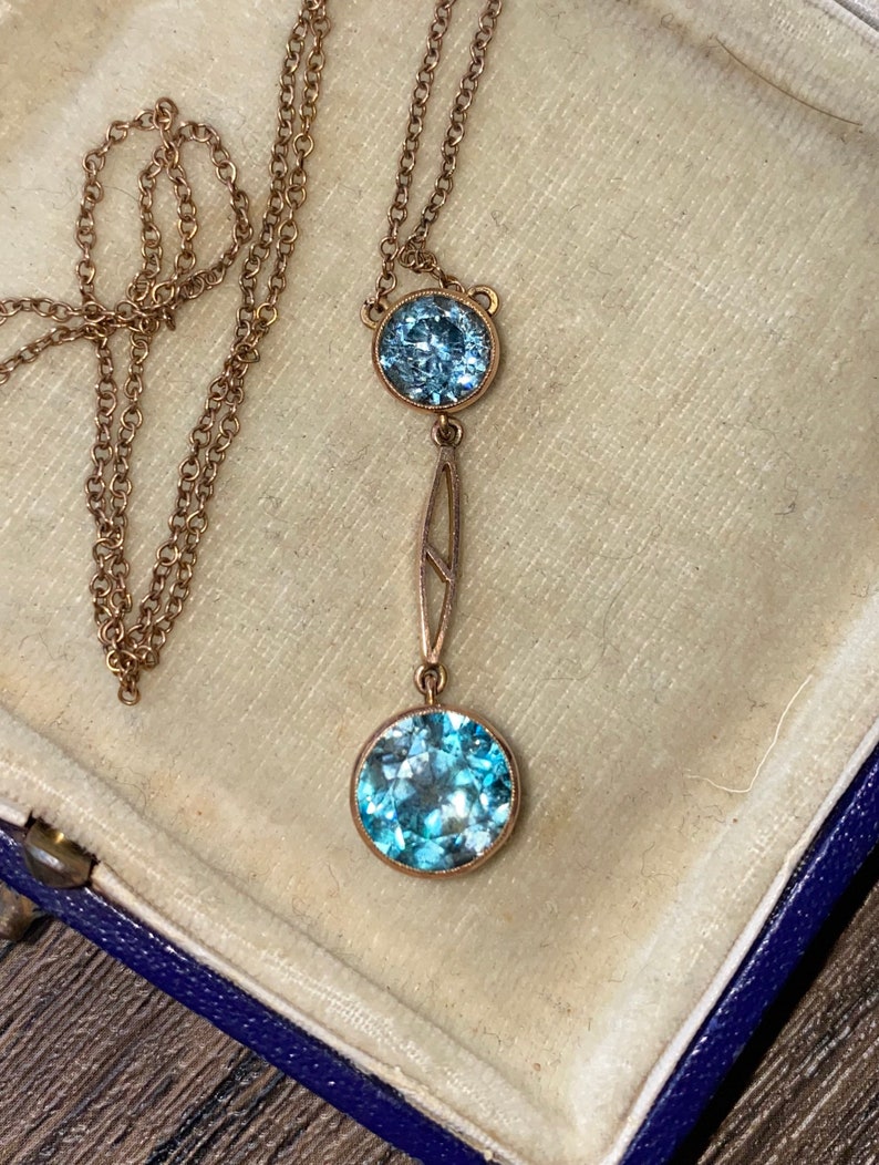 Op de afbeelding: Een gouden ketting met een hanger met twee ronde, lichtblauwe edelstenen. De hanger heeft een delicaat art deco-ontwerp.