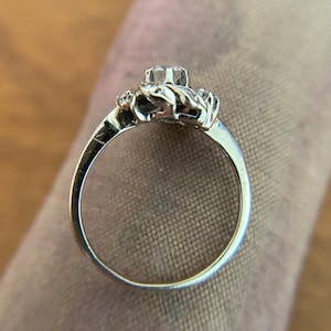 Puede incluir: Un anillo de plata con una gran piedra preciosa transparente rodeada de piedras preciosas m&aacute;s peque&ntilde;as. El anillo est&aacute; engastado en un delicado dise&ntilde;o floral.