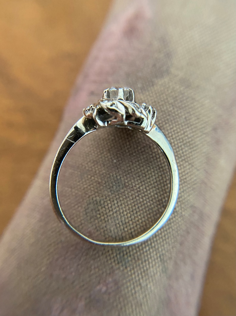 Puede incluir: Un anillo de plata con una gran piedra preciosa transparente rodeada de piedras preciosas m&aacute;s peque&ntilde;as. El anillo est&aacute; engastado en un delicado dise&ntilde;o floral.