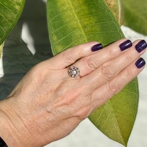 Puede incluir: Un anillo de plata con una piedra grande, redonda y transparente rodeada de piedras m&aacute;s peque&ntilde;as. El anillo est&aacute; en un dedo.