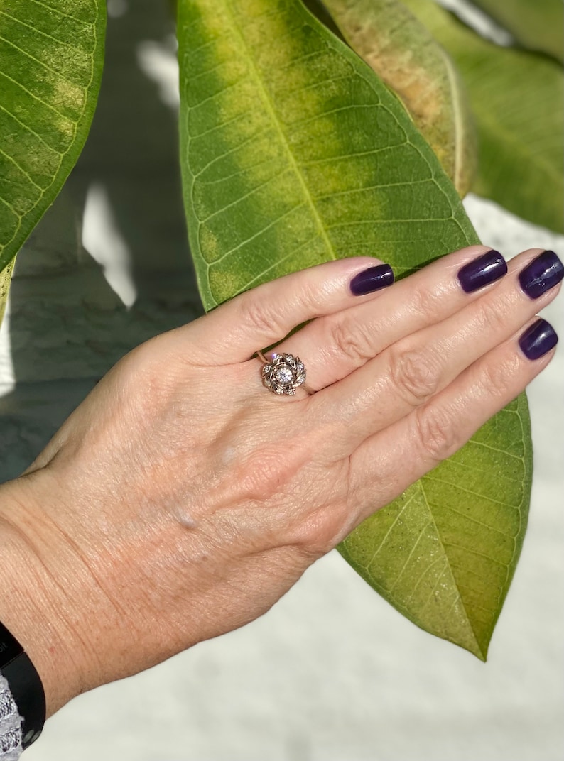 Puede incluir: Un anillo de plata con una piedra grande, redonda y transparente rodeada de piedras m&aacute;s peque&ntilde;as. El anillo est&aacute; en un dedo.