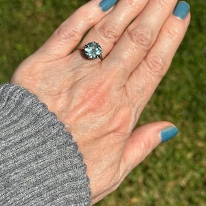 Op de afbeelding: Een zilveren ring met een grote, ronde, lichtblauwe edelsteen in een bezel-setting. De ring zit aan een vinger.