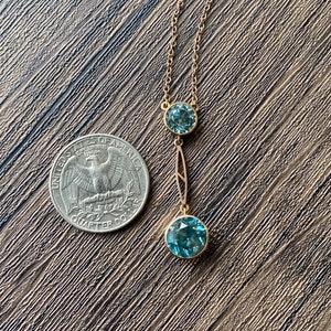 Op de afbeelding: Een gouden ketting met twee blauwe edelsteen hangers. De grotere hanger bevindt zich aan de onderkant van de ketting en de kleinere hanger erboven. De ketting ligt op een bruin houten oppervlak.
