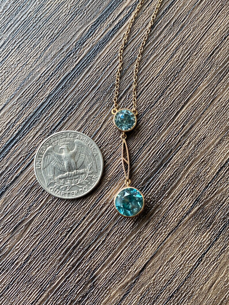 Op de afbeelding: Een gouden ketting met twee blauwe edelsteen hangers. De grotere hanger bevindt zich aan de onderkant van de ketting en de kleinere hanger erboven. De ketting ligt op een bruin houten oppervlak.