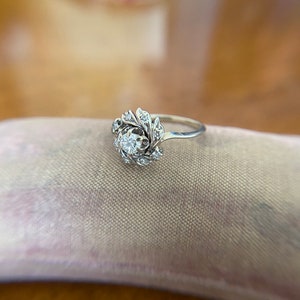Puede incluir: Un anillo de plata con un diamante redondo y claro en el centro, rodeado de diamantes m&aacute;s peque&ntilde;os en un dise&ntilde;o floral.