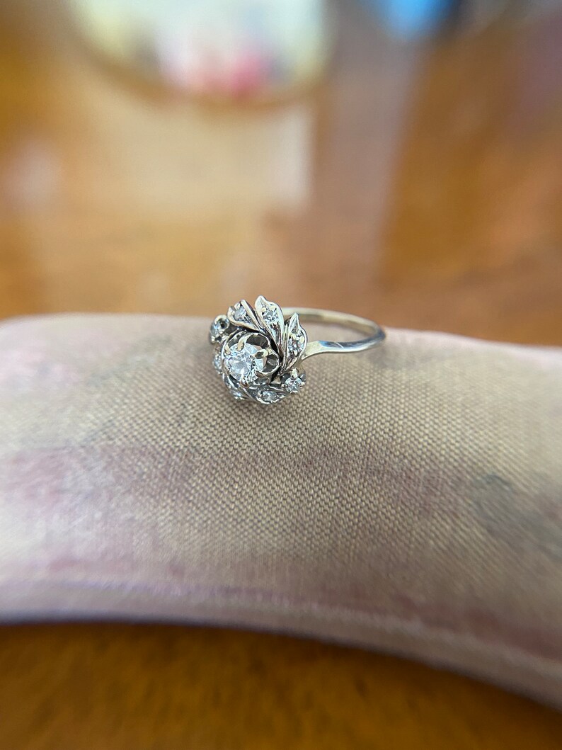 Puede incluir: Un anillo de plata con un diamante redondo y claro en el centro, rodeado de diamantes m&aacute;s peque&ntilde;os en un dise&ntilde;o floral.