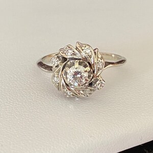 Puede incluir: Un anillo de plata con un gran diamante redondo en el centro, rodeado de diamantes m&aacute;s peque&ntilde;os engastados en un dise&ntilde;o floral.
