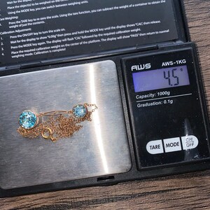 Op de afbeelding: Een gouden ketting met drie lichtblauwe edelstenen op een digitale weegschaal die 4,5 gram aangeeft. De weegschaal is gemarkeerd met "AWS-1KG" en heeft een capaciteit van 1000 gram met een graduatie van 0,1 gram.