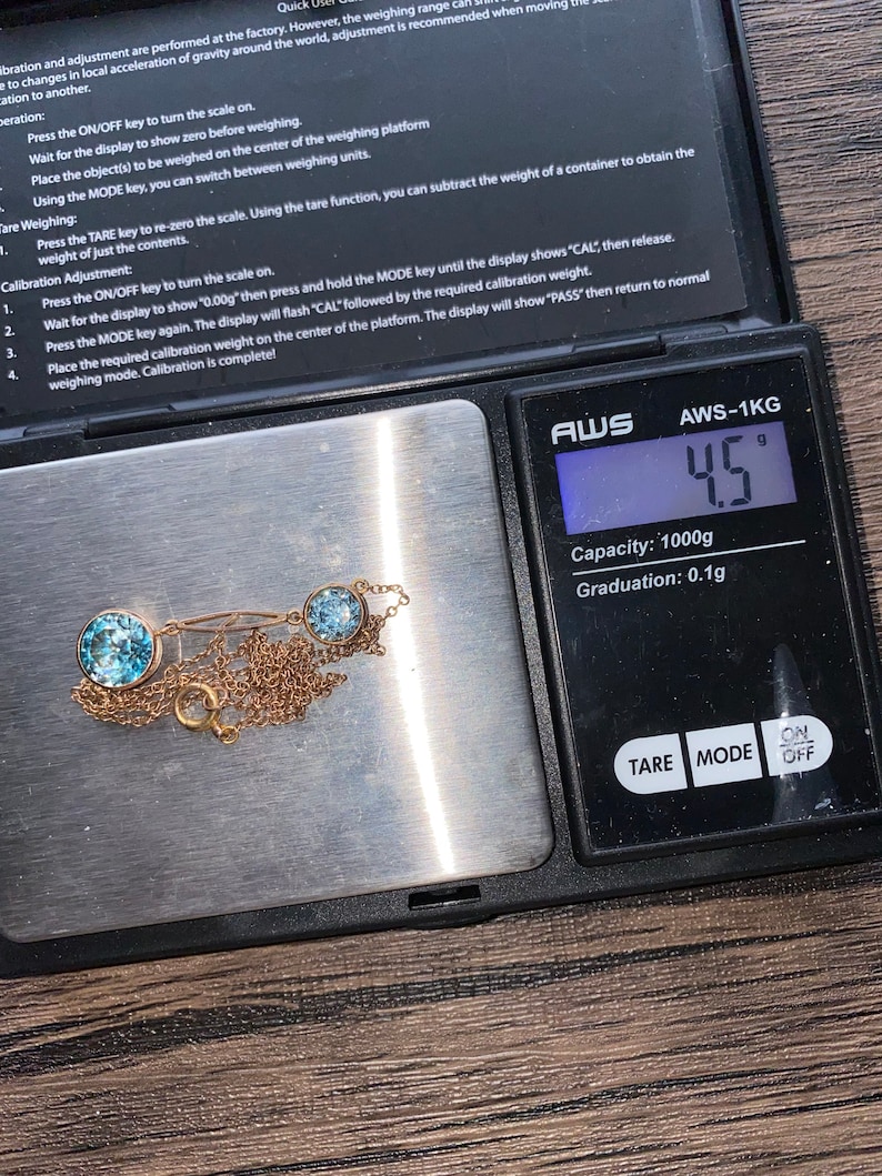 Op de afbeelding: Een gouden ketting met drie lichtblauwe edelstenen op een digitale weegschaal die 4,5 gram aangeeft. De weegschaal is gemarkeerd met "AWS-1KG" en heeft een capaciteit van 1000 gram met een graduatie van 0,1 gram.