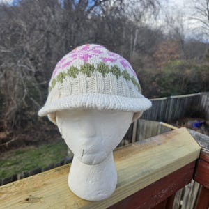 Puede incluir: Un gorro de punto color crema con ala, con un patrón geométrico en rosa, verde y naranja. El gorro se exhibe en una cabeza de maniquí blanca, mostrando su diseño y textura. El gorro es un accesorio de invierno.