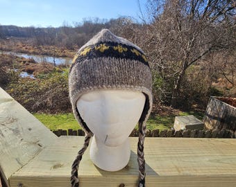 Pode incluir: Um gorro de inverno tricotado à mão com protetores de orelha. O chapéu é principalmente cinza com uma faixa de padrões geométricos pretos e amarelos. Tassels trançados pendem dos protetores de orelha. O chapéu é exibido em uma cabeça de manequim branca.