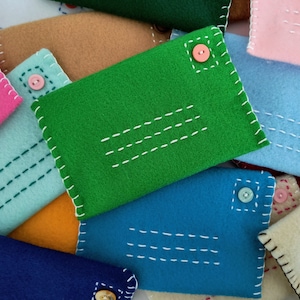 May include: A collection of felt envelopes in various colors, including green, blue, pink, and beige. Each envelope has a button closure and decorative stitching.
