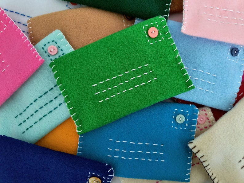 May include: A collection of felt envelopes in various colors, including green, blue, pink, and beige. Each envelope has a button closure and decorative stitching.