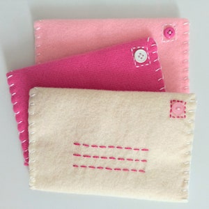 May include: Three felt envelopes in pink, fuchsia, and cream with pink buttons and white stitching. The cream envelope has a pink stitched rectangle in the center.