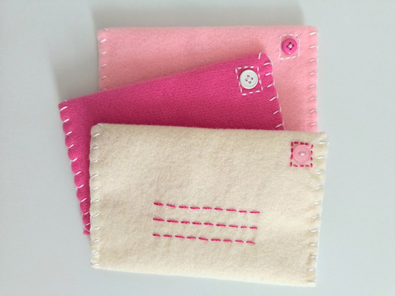 May include: Three felt envelopes in pink, fuchsia, and cream with pink buttons and white stitching. The cream envelope has a pink stitched rectangle in the center.