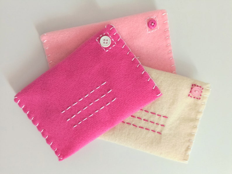 May include: Three felt envelopes in pink, light pink, and cream with white and pink stitching. Each envelope has a button closure.
