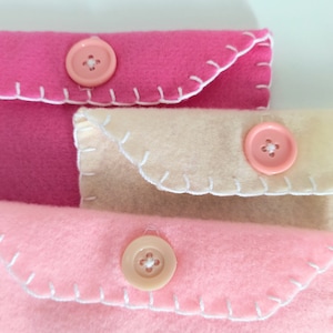 May include: Three felt fabric envelopes with white stitching and pink buttons. The envelopes are stacked on top of each other, with the top envelope being pink, the middle envelope being beige, and the bottom envelope being pink.