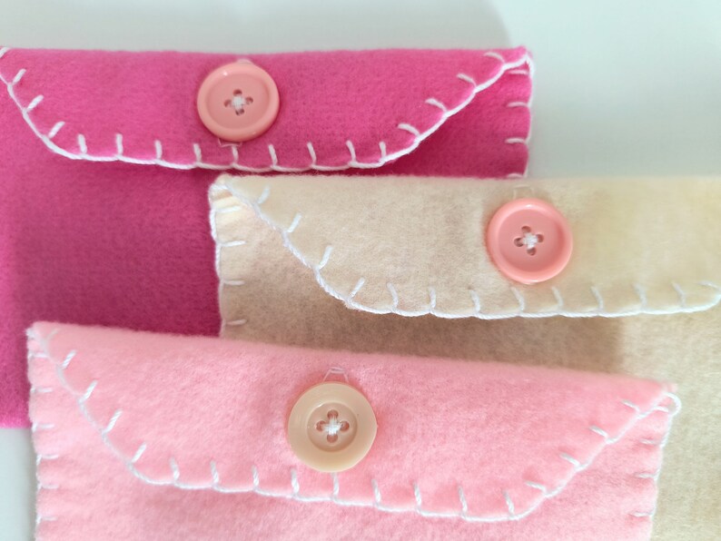 May include: Three felt fabric envelopes with white stitching and pink buttons. The envelopes are stacked on top of each other, with the top envelope being pink, the middle envelope being beige, and the bottom envelope being pink.