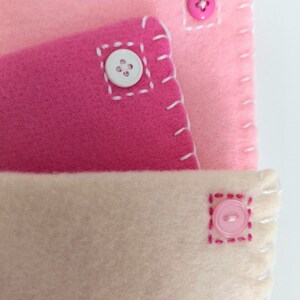 May include: Three layers of fabric in pink, light pink, and cream colors, each with a button sewn on. The buttons are white, pink, and pink. The fabric is stitched with pink thread.