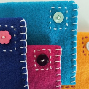 May include: Four fabric pouches with different colored felt and contrasting stitching. Each pouch has a button sewn on the front. The buttons are pink, green, black, and white.
