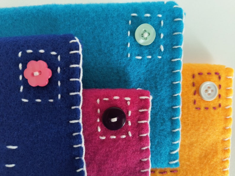 May include: Four fabric pouches with different colored felt and contrasting stitching. Each pouch has a button sewn on the front. The buttons are pink, green, black, and white.