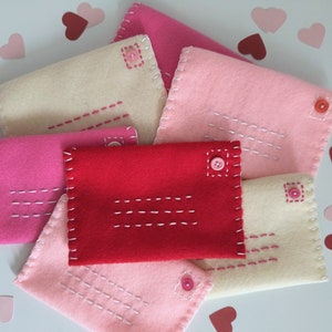 May include: A collection of handmade felt envelopes in shades of pink, red, and cream. Each envelope features white stitching details and a button closure. Scattered around are paper heart cutouts in coordinating colors.
