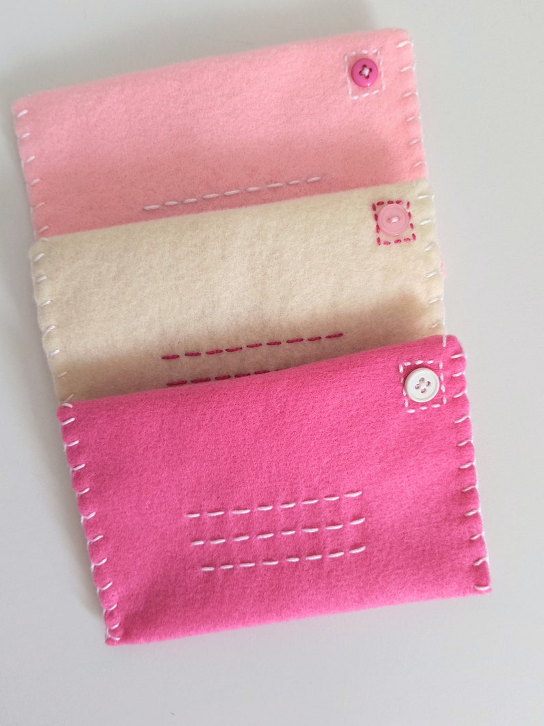 May include: Three felt envelopes in pink, cream, and hot pink with white stitching and pink buttons.