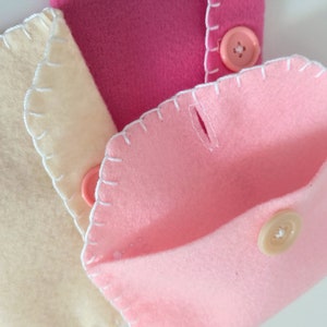 May include: Three felt pouches with white stitching and button closures. The pouches are in shades of pink and cream.
