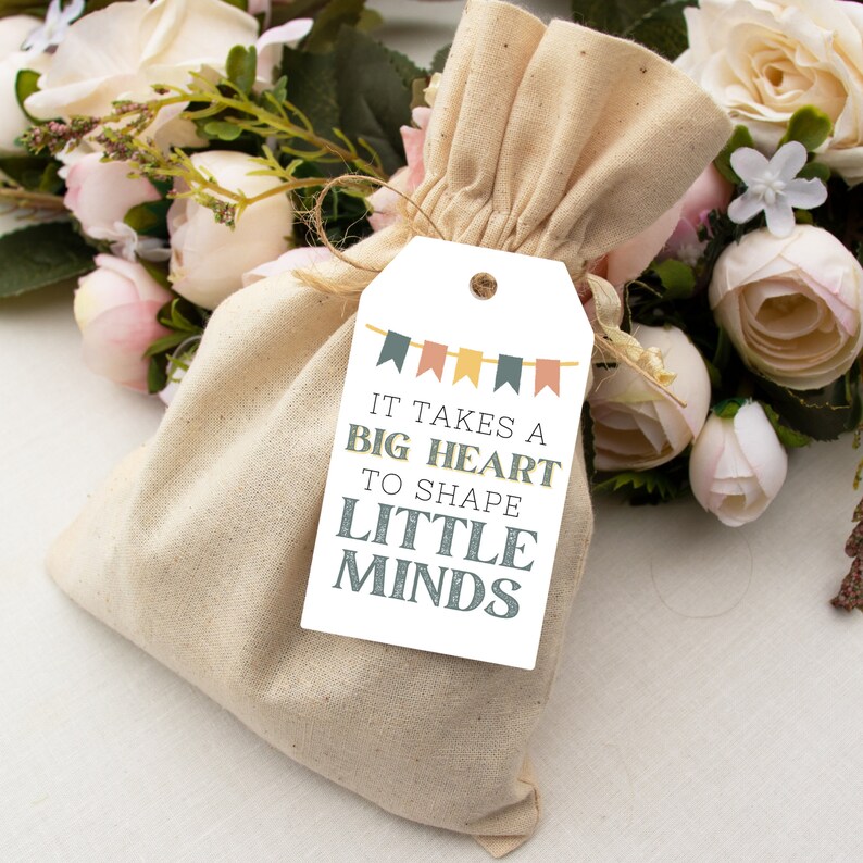 May include: A white gift tag with the text "It takes a big heart to shape little minds". The tag is tied to a small, beige fabric bag with a drawstring. The bag is surrounded by pink and white flowers.