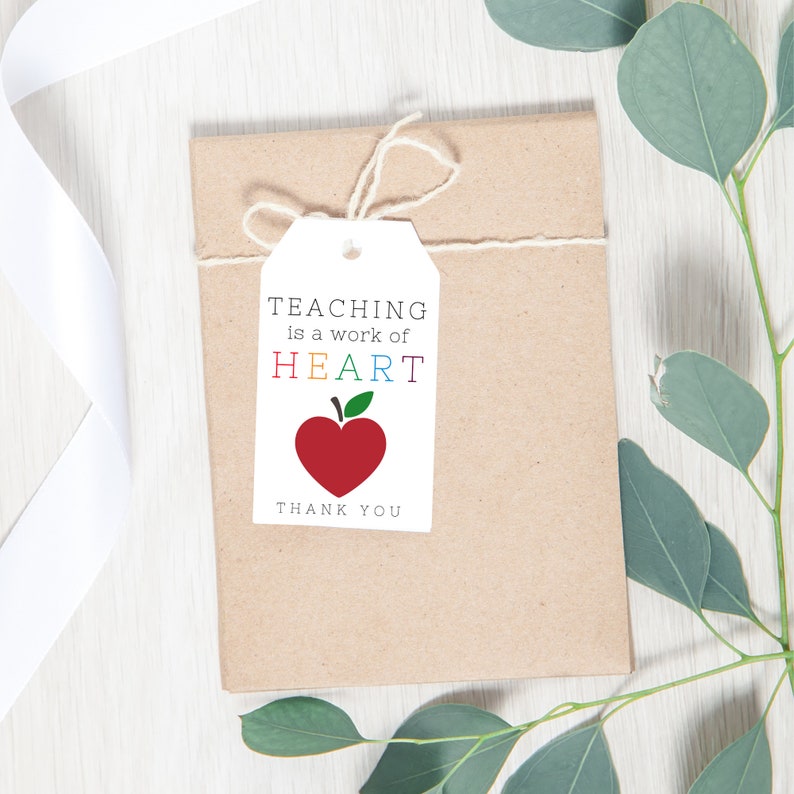 May include: A white gift tag with the text "TEACHING is a work of HEART" in rainbow colors and a red apple heart. The tag is tied with twine to a brown paper gift bag.  Thank you.