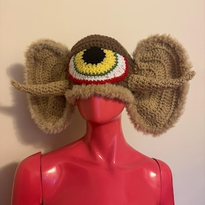 May include: A whimsical, handmade hat featuring a large, central eye design with brown, yellow, red, and white details. The hat has fluffy, tan ear flaps and a faux fur trim, creating a unique and playful accessory.