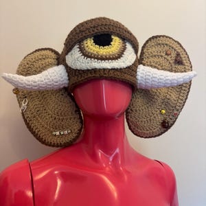 May include: A brown crocheted hat with a large eye design, featuring a black pupil, yellow iris, and white accents. The hat has two tan ear-shaped extensions with white horn-like details and decorative beads. The word "WEIRD" is spelled out on one ear.