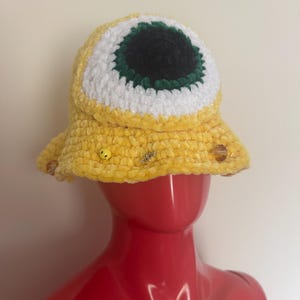 May include: A yellow crochet bucket hat with a large eye design. The eye features a black pupil, white iris, and green outline. The hat is decorated with small embellishments and is displayed on a red mannequin head.