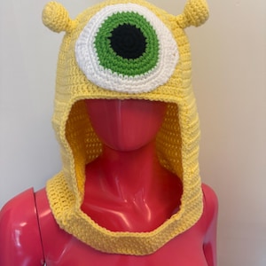 May include: A yellow crocheted hood with a large eye design. The eye has a white iris, a green outer ring, and a black pupil. The hood features two small, rounded antennae. Displayed on a mannequin.