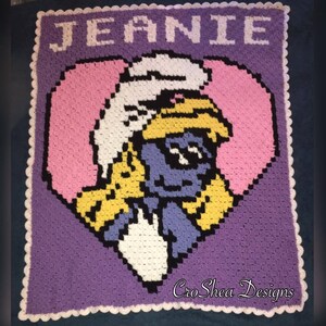 May include: A purple crocheted blanket with a white border. The blanket features a heart-shaped design with a cartoon character inside. The character is blue with a yellow hat and white hair. The text "JEANIE" is at the top of the blanket and "CroShea Designs" is at the bottom.