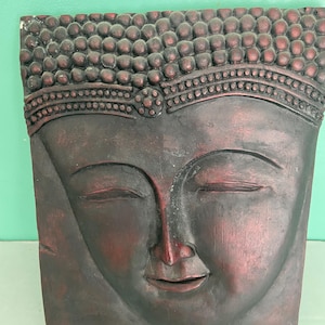 May include: A square, wooden wall hanging featuring a carved Buddha face. The face is a dark reddish-brown color with closed eyes and a serene expression. The top of the head has a textured pattern of small bumps.