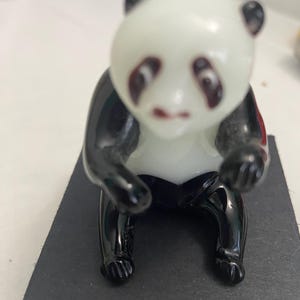 May include: A small, glass panda figurine with a white head and body, black arms, legs, and ears. The panda is sitting on a black rectangular base. The figurine has a glossy finish.