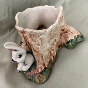 May include: A ceramic planter shaped like a tree stump with a white rabbit peeking out from the side. The planter is painted in shades of brown, green, and white.